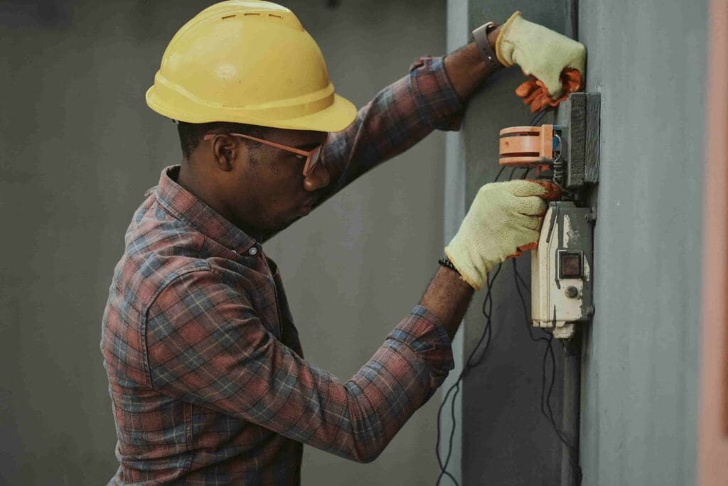 A skilled electrician wearing a yellow hard hat and safety gloves working diligently on an electrical panel, representing the diligence and expertise The People Biz brings to HR for small businesses. This image symbolizes the professional and precise approach needed in managing HR functions to enhance productivity and growth in small business settings.